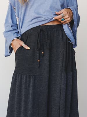 A close-up mid-shot of a charcoal tiered maxi skirt styled with a relaxed periwinkle blue top, accessorized with stacked silver rings and a beaded bracelet — the kind of effortless pairing that looks intentional without trying too hard.
