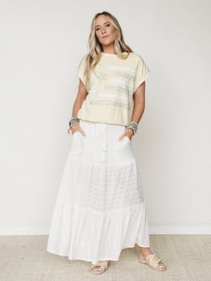 A breezy, sun-washed look — cream tiered maxi skirt with a textured stripe detail paired with a butter yellow graphic tee, stacked turquoise bracelets, and slide sandals, shot clean against a soft white studio wall.