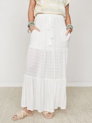 A white tiered maxi skirt with a drawstring tassel waist and lace panel detail, styled with a soft yellow embroidered top, stacked beaded bracelets, and raffia sandals — the kind of outfit that looks like a slow morning somewhere warm.