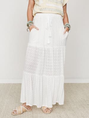 A white tiered maxi skirt with a drawstring tassel waist and lace panel detail, styled with a soft yellow embroidered top, stacked beaded bracelets, and raffia sandals — the kind of outfit that looks like a slow morning somewhere warm.