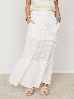 A cream tiered maxi skirt with subtle woven texture and a lace-panel inset is styled with beaded bracelets, a pale yellow top, and woven sandals — effortlessly relaxed and ready for somewhere warm.