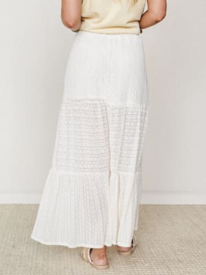 A cream tiered maxi skirt shot from behind, showing off its textured fabric, sheer lace inset panel, and ruffled hem — soft, easy, and made for warm-weather dressing.