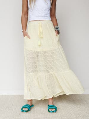 A breezy butter-yellow tiered maxi skirt with a sheer lace panel and tassel drawstring waist, styled with a white eyelet crop top and teal crochet sandals — the kind of outfit that looks like it was assembled effortlessly on a warm morning.