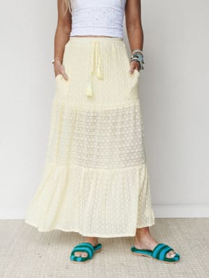 A soft butter-yellow tiered maxi skirt with a lace inset panel and tassel drawstring waist, styled with a white eyelet crop top and teal crochet sandals — easy, sunny, and just the right amount of boho.
