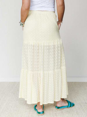 A soft butter-yellow tiered maxi skirt shot from the back, styled with a white eyelet crop top and teal crochet sandals — easy, warm-weather dressing with a relaxed boho feel.