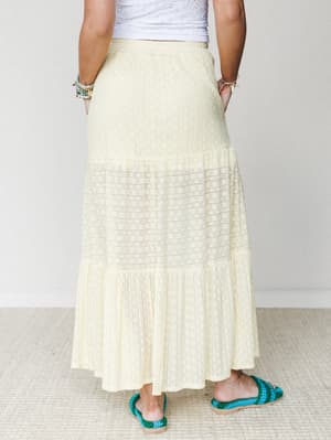A soft butter-yellow tiered maxi skirt shot from the back, styled with a white eyelet crop top and teal crochet sandals — easy, warm-weather dressing with a relaxed boho feel.