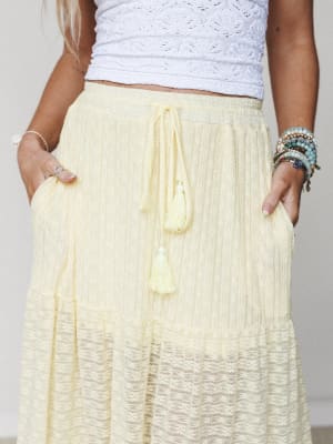 A close-up torso shot of a butter yellow tiered maxi skirt with lace hem detail and tassel drawstring, paired with a white crochet crop top and stacked boho bracelets — the kind of outfit that makes a farmers market feel like a little occasion.