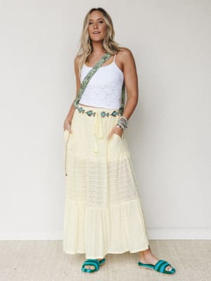 A sun-warmed look built around a soft butter-yellow tiered maxi skirt, styled with a white eyelet crop cami, teal crochet sandals, and a floral crossbody strap — the kind of outfit that makes a regular afternoon feel like a trip somewhere warm.