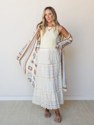 A breezy, all-white moment — cropped eyelet top paired with a tiered lace maxi skirt and held open against a granny square crochet kimono in soft pastels, giving off that effortless something-borrowed, something-boho energy.