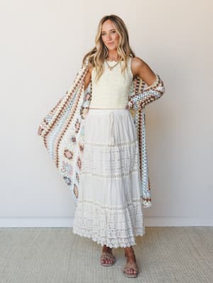A breezy, all-white moment — cropped eyelet top paired with a tiered lace maxi skirt and held open against a granny square crochet kimono in soft pastels, giving off that effortless something-borrowed, something-boho energy.