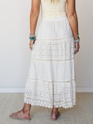A back-view shot of a tiered ivory maxi skirt with alternating lace panels and eyelet trim, styled with a cream textured top, stacked turquoise bracelets, and flat sandals — soft, romantic, and made for somewhere warm.