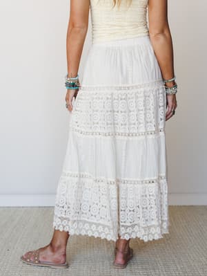 A back-view shot of a tiered ivory maxi skirt with alternating lace panels and eyelet trim, styled with a cream textured top, stacked turquoise bracelets, and flat sandals — soft, romantic, and made for somewhere warm.