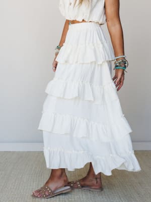 A tiered ivory maxi skirt with ruffled layers and a smocked waistband, styled with a cropped cream top, stacked turquoise bracelets, and tan studded sandals — the kind of all-white outfit that feels effortless rather than overdressed.