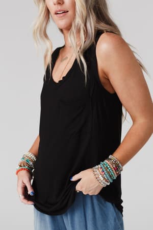 A woman with wavy blonde hair wears The Nest Shelli Scoop Neck Tank Top in black, styled with light blue jeans and colorful beaded bracelets on both wrists. She poses against a plain light background.