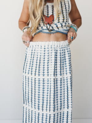 A crochet two-piece set in ivory and dusty blue — cropped top and maxi skirt with an open-weave texture — styled with layered turquoise and mixed-metal jewelry and a red butterfly pendant necklace for a free-spirited, collected feel.
