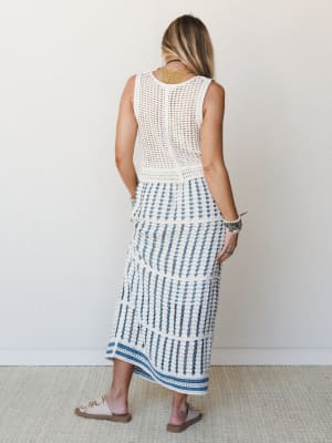 Shot from behind, a model wears a cream open-knit sleeveless top and matching crochet midi skirt with blue stripe detailing at the hem — the kind of set that feels equally at home at a seaside lunch or a slow summer afternoon.