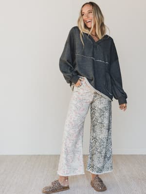 Woman wearing a dark hoodie and patterned pants standing against a white wall.