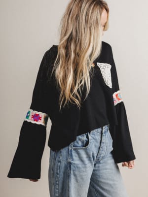 Woman wearing a black boho blouse with colorful sleeve designs and blue jeans on a neutral background