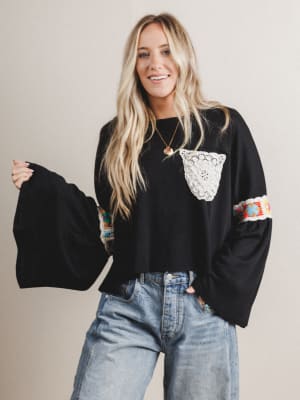 Woman wearing a black boho top with colorful sleeves and a lace pocket, standing against a plain background.