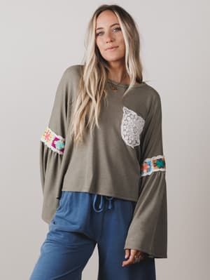 Woman wearing an olive green long-sleeve top with colorful accents and blue pants against a plain background