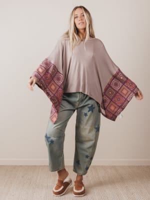 Woman wearing a beige poncho with patterned edges and blue jeans against a plain background