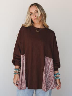 A relaxed, oversized ribbed top in deep chocolate brown with contrasting floral and tribal-print patchwork panels at the sides and cuffs, styled with light blue jeans and stacked turquoise and beaded bracelets — effortlessly lived-in with a boho edge