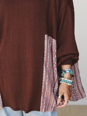 A close-up detail shot of a rust-brown ribbed tunic top with contrast patchwork side panels in a dusty rose tribal print, styled with a stacked bracelet set including a turquoise 'MAMA' letter bracelet and beaded strands — cozy and personal all at on