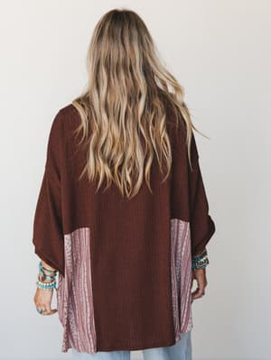 Shot from the back, a model in a relaxed oversized patchwork top layers rich cocoa brown textured fabric with dusty rose printed side panels — boho layering that feels intentional, not try-hard, finished with stacked turquoise and beaded bracelets.