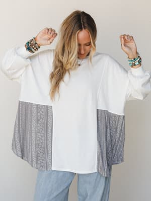 An oversized mixed-fabric pullover top in off white with grey floral and geometric patchwork panels on the sides, styled with light wash wide-leg jeans and stacked boho bracelets — the kind of top that looks intentional without trying.