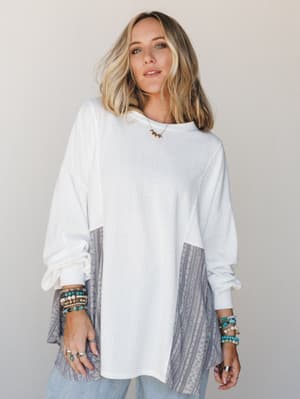 An oversized long-sleeve top in off-white with contrast tribal-stripe patchwork panels at the sides and lower sleeves, styled with stacked turquoise and beaded bracelets, silver rings, and a dainty gold necklace — easy and expressive, the kind of top