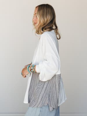 A side-profile shot of a model in a relaxed patchwork top — white textured fabric up top, transitioning into a grey geometric stripe panel at the hem — styled with stacked turquoise bracelets and light-wash jeans for an effortlessly personal, boho-ca