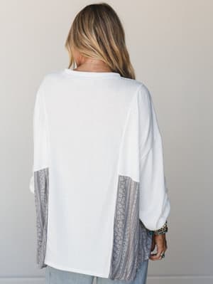 A back-facing shot of a relaxed, oversized knit top in off-white with contrast patchwork side panels in a grey floral-stripe print, styled with light wash jeans and layered boho bracelets — the kind of top that looks intentional without trying too ha