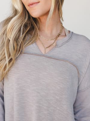 A close-up of a soft, dusty lavender-brown pullover with exposed seam detailing at the shoulders, styled with layered gold chain necklaces — the kind of top that looks effortless whether you're on a coffee run or working from the couch.