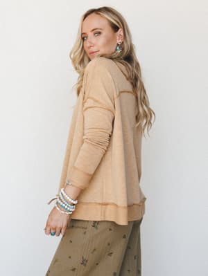 A relaxed long-sleeve pullover in warm sandy mustard is shot from the back at three-quarters, paired with an olive floral skirt and stacked turquoise bracelets — the kind of effortless, collected look that feels just as good as it looks.
