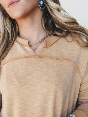 A close-up detail shot of a warm mustard pullover with contrast stitching and a notched neckline, styled with layered gold chain necklaces and turquoise statement earrings for an effortless Western-boho feel.