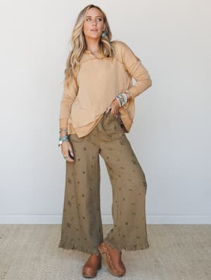 A relaxed, earthy outfit pairing a soft mustard long-sleeve pullover with wide-leg olive floral pants and brown clogs — the kind of effortless combo that looks like it came together on its own.