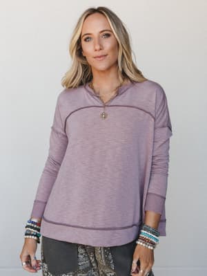 A relaxed mauve pullover with exposed seam detailing worn over a paisley bottom, styled with layered gold necklaces and stacks of beaded bracelets — the kind of top that looks intentional even on a Tuesday.