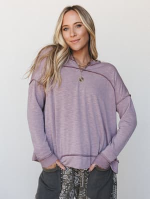 A relaxed, oversized pullover hoodie in a soft washed mauve worn with paisley pants and layered gold necklaces — the kind of outfit that looks effortless because it actually is.
