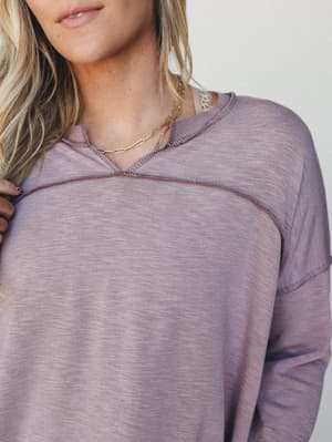 A close-up of a dusty mauve pullover with a lived-in, burnout texture and exposed contrast stitching — the kind of top that looks equally good with jeans or pajama pants, layered with a gold chain necklace.