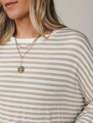 A close-up lifestyle shot of a relaxed long-sleeve striped top in warm tan and cream, styled with layered gold chain necklaces — the kind of effortless everyday piece that feels intentional without trying too hard.
