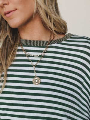 A close-up lifestyle shot of a green and white striped tee with a contrast ribbed neckline, styled with a layered gold paperclip chain necklace featuring a coin pendant — easy, expressive, and the kind of outfit that looks effortless without trying.