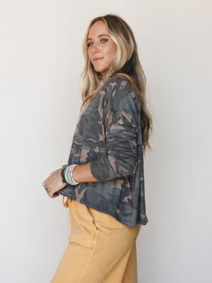 A relaxed side profile shot of a long-sleeve camo top paired with golden mustard wide-leg pants, styled with stacked beaded bracelets — the kind of effortless, lived-in look that feels intentional without trying too hard.