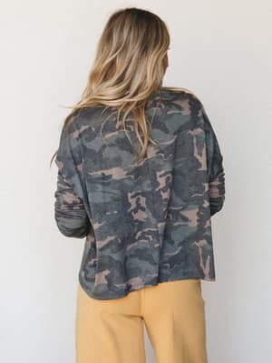 Shot from behind, a model wears an oversized camo long sleeve top in muted army tones paired with warm golden joggers — effortless and lived-in, the kind of outfit that makes staying in feel intentional.