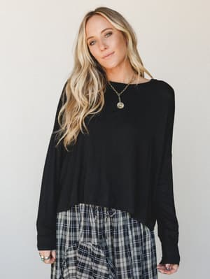 A relaxed black long sleeve top styled with a plaid skirt and gold pendant necklace — effortless and lived-in, the kind of outfit that works as hard as she does without looking like it's trying.
