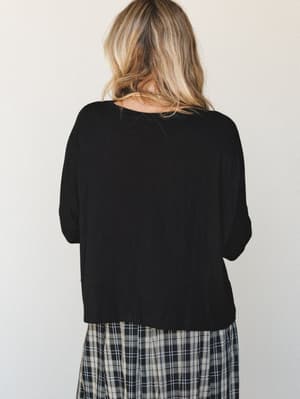 A relaxed black long sleeve top paired with a plaid skirt, shot from the back against a soft neutral studio wall — easy, unfussy, the kind of top that goes with everything and asks nothing of you.