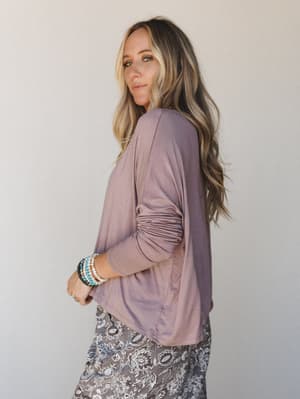 A relaxed long sleeve top in a soft mushroom mauve, shot in profile to show the ruched sleeve detail — styled with a grey floral print bottom and stacked boho bracelets for an effortlessly put-together everyday look.
