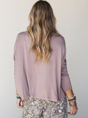 A relaxed, back-facing shot of a model in a soft mauve long-sleeve top paired with floral print pants and stacked boho bracelets — effortless and lived-in, like a Sunday that actually went right.