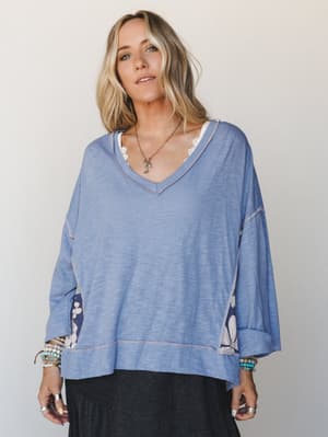 A relaxed, oversized v-neck top in soft periwinkle blue with floral contrast panels peeking out at the sides and sleeves, layered over a lace-trim cami and styled with stacked beaded bracelets and a delicate pendant necklace — the kind of top that lo