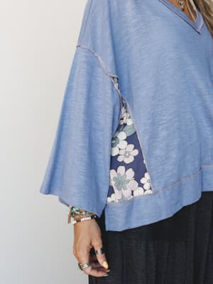 A close-up detail shot of a soft periwinkle blue top with bell sleeves and exposed seam stitching, layered over a navy floral dress — styled with stacked beaded bracelets, silver rings, and a turquoise necklace for that collected, lived-in boho feel.