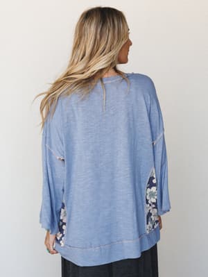 Shot from the back, a relaxed oversized top in soft periwinkle blue shows off its floral contrast panels at the sides and bell sleeves — the kind of top that feels like a deep exhale.